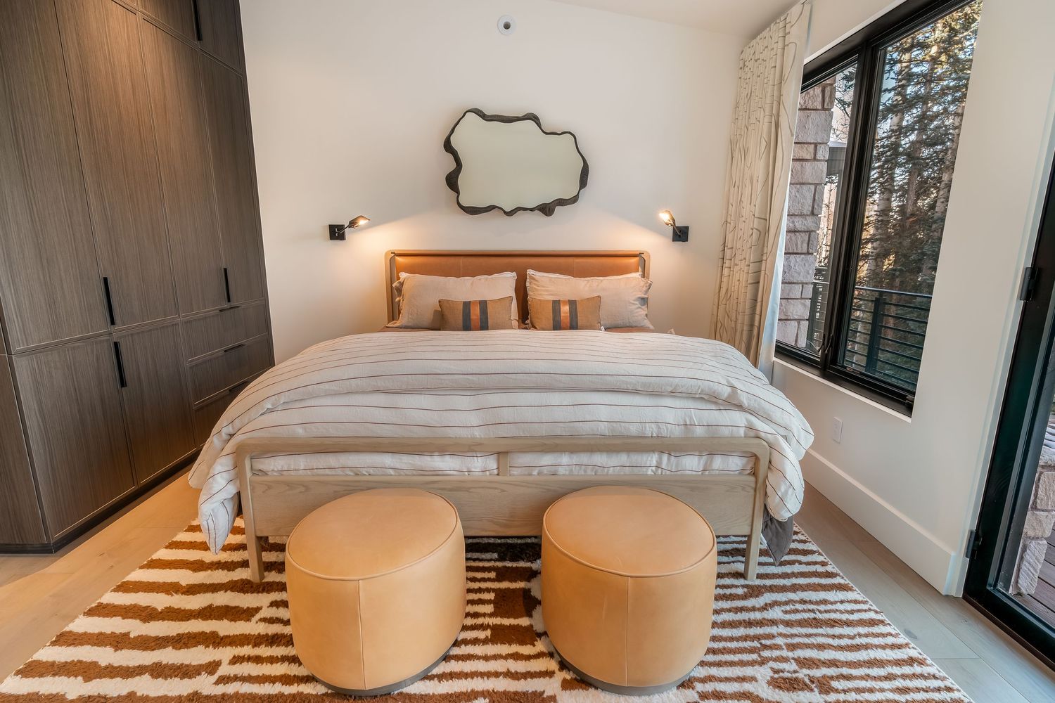 Modern bedroom interior with white bedding, striped rug, and natural light from large windows.