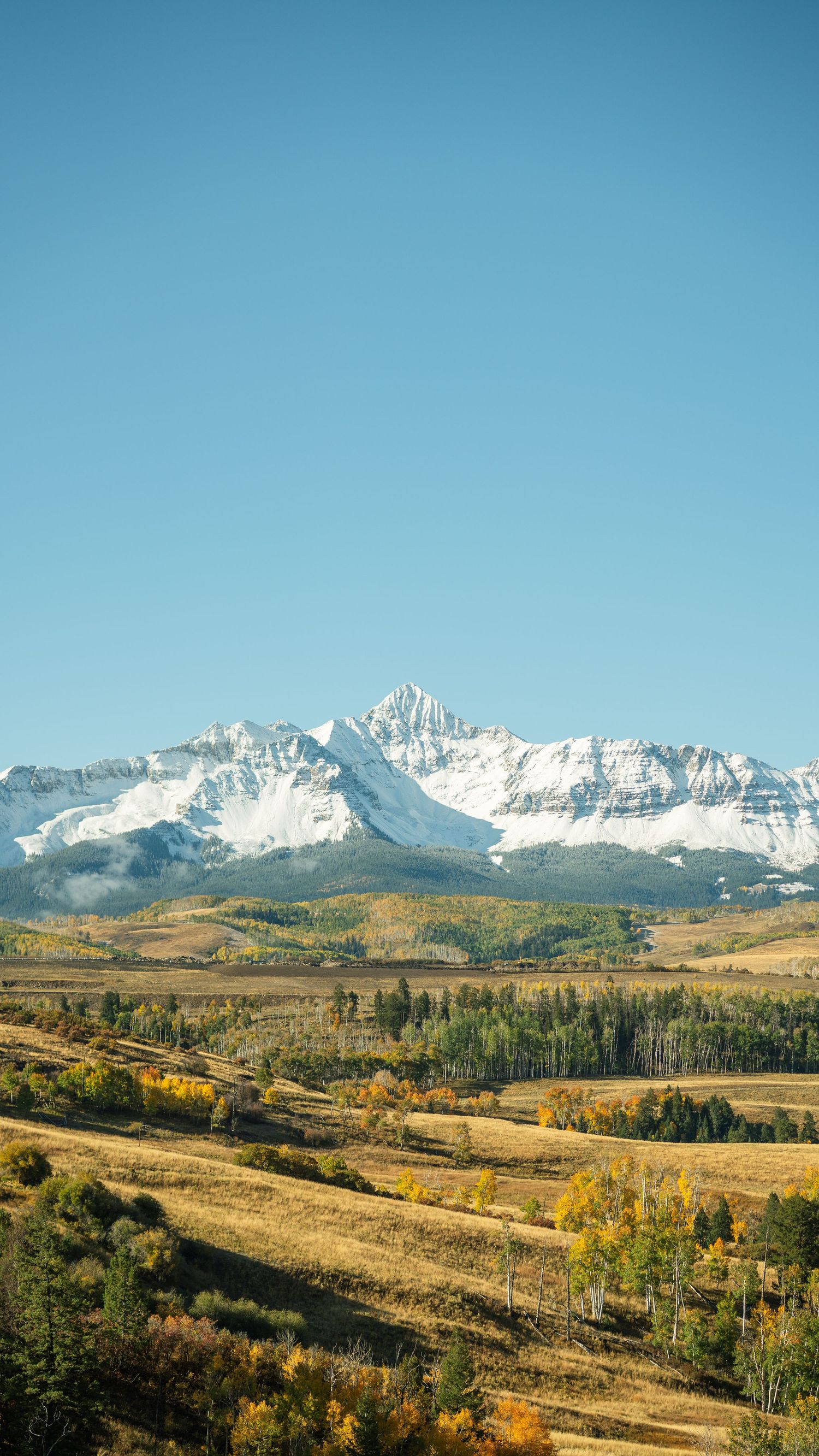 Snow-capped mountain range rises above golden autumn valleys and rolling hills.