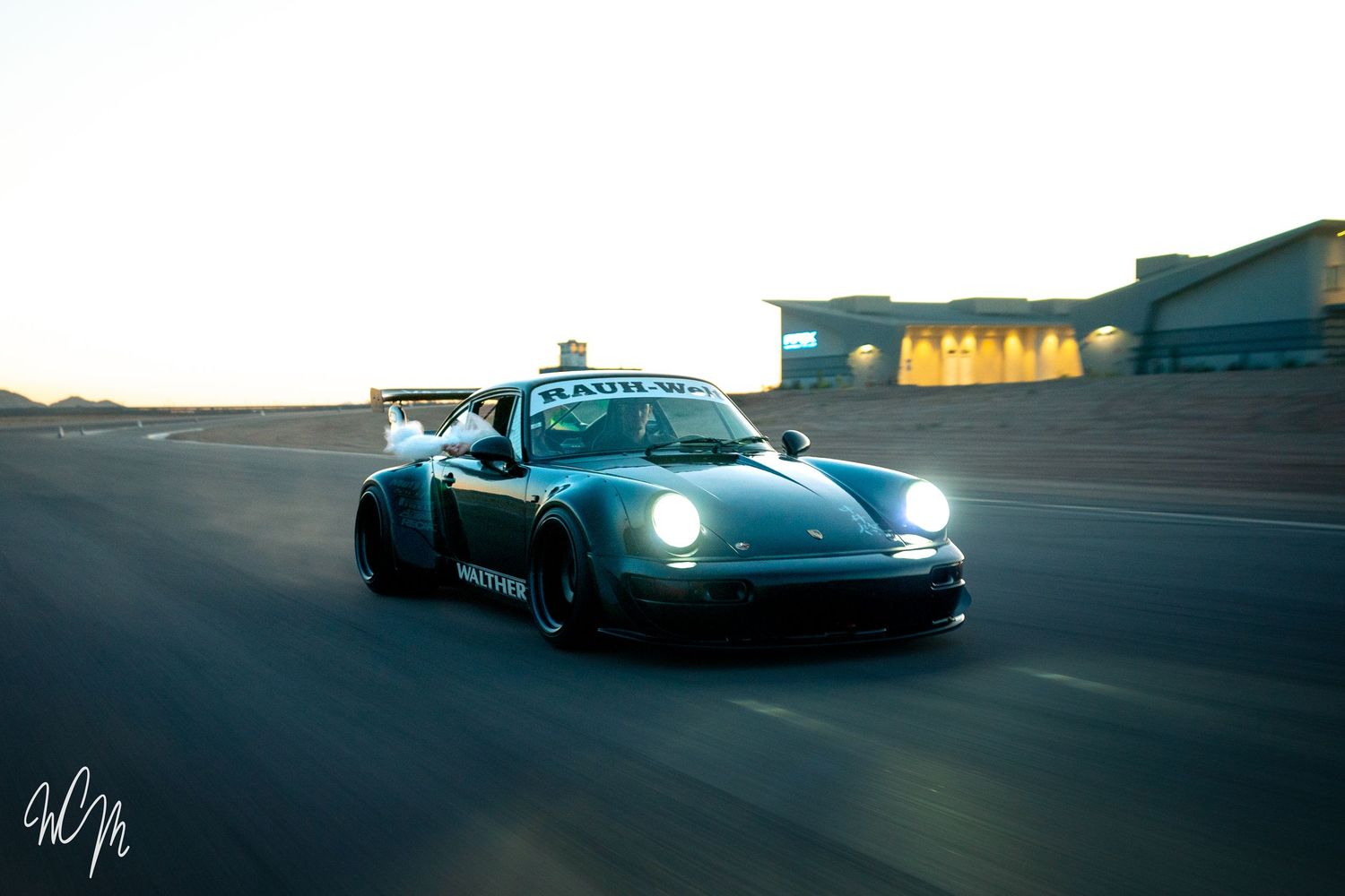 Modified blue Porsche 911 racing on track with headlights illuminated at dusk.