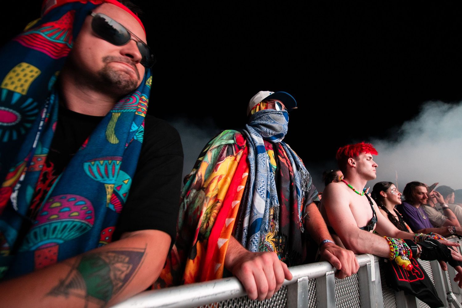 Colorful festival crowd at a concert, featuring fans in vibrant outfits and face coverings leaning on a barrier.