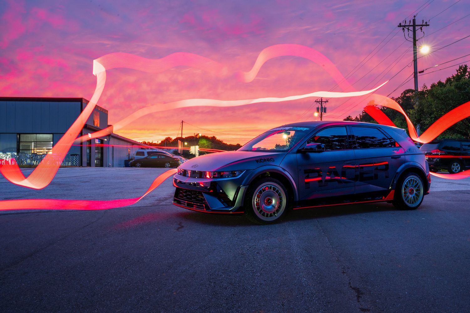 Modified sports car with light trails at sunset in a dramatic pink and purple sky.