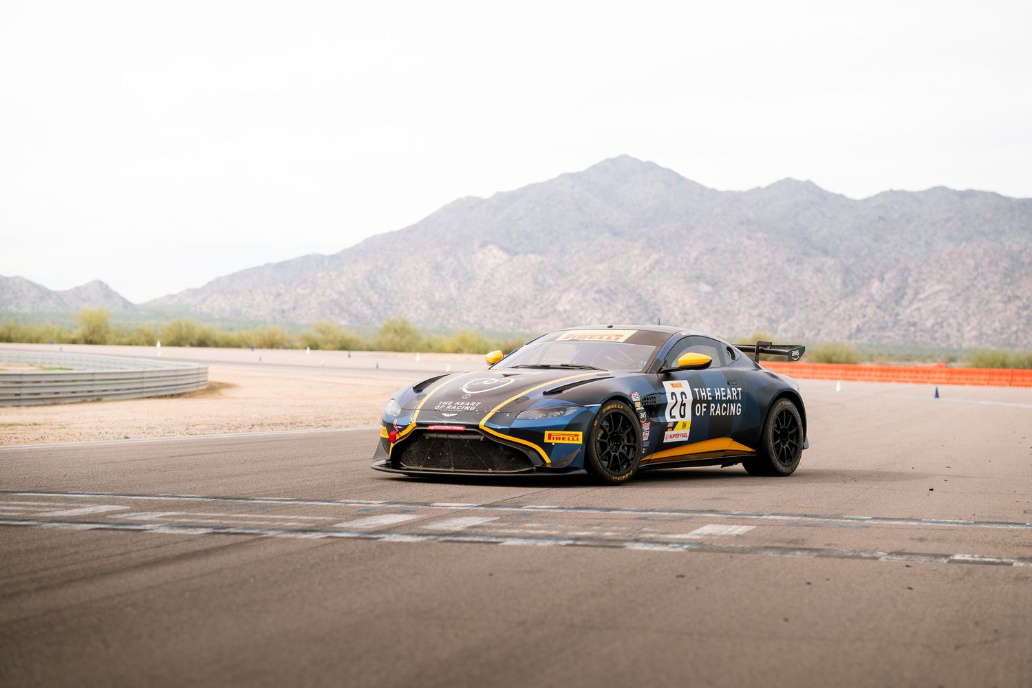 A sleek Aston Martin racing car with black and orange livery sits on a desert track with mountains in the background.
