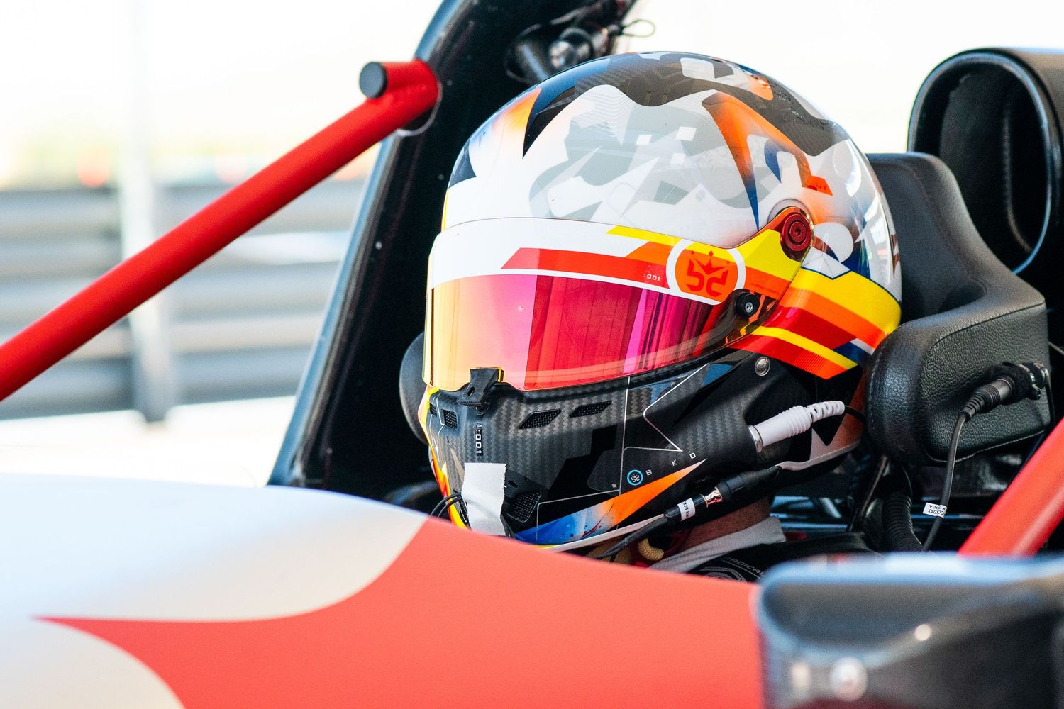 Race car cockpit view showing helmet with red visor and roll cage structure.