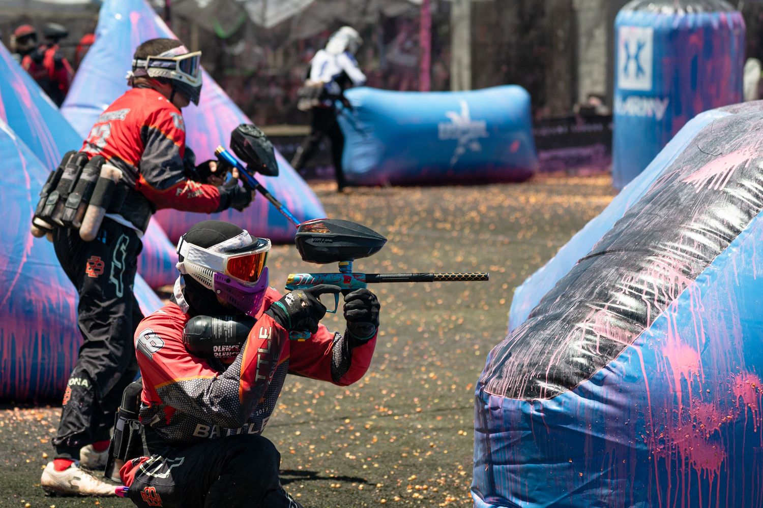 Two paintball players in red gear take cover behind inflatable bunkers during a competitive speedball match.