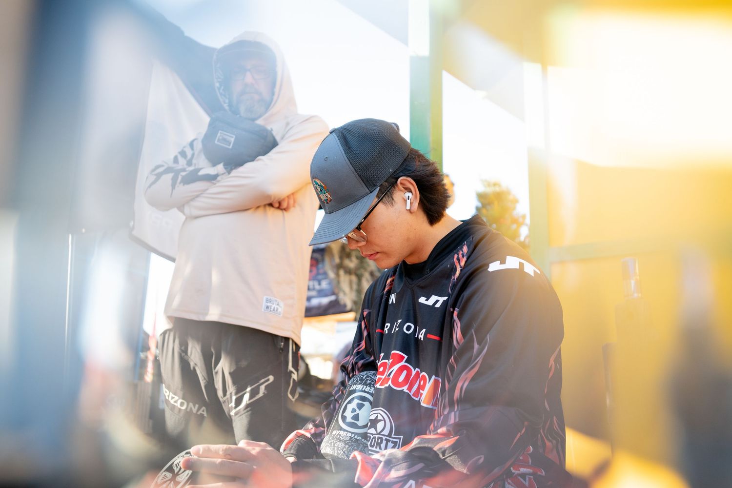 Two players in paintball gear prepare under warm sunlight at an outdoor paintball tournament.