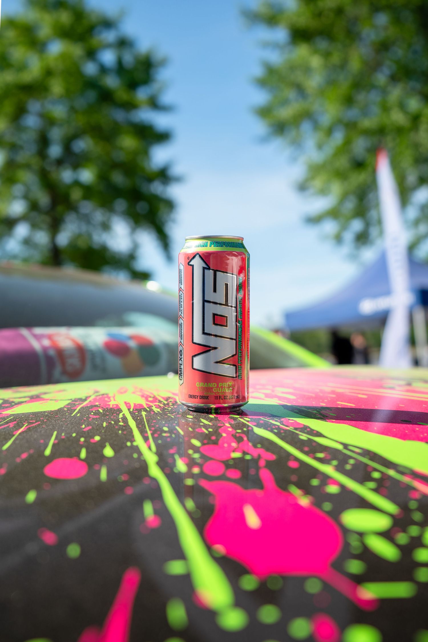 NOS energy drink can balanced on colorful neon splatter paint car hood at outdoor automotive event.