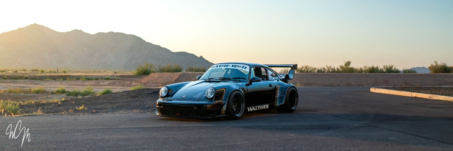A blue Porsche 911 RWB sits on a desert road with mountains in the background during sunset.