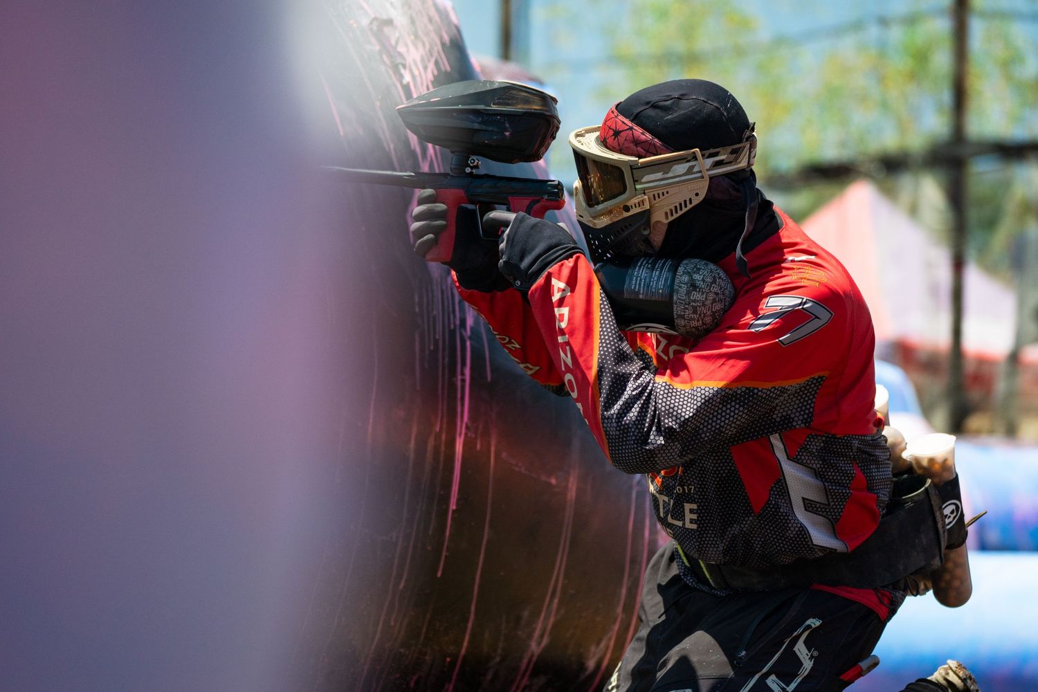 A paintball player in red gear crouches behind a bunker during an outdoor competitive paintball tournament.