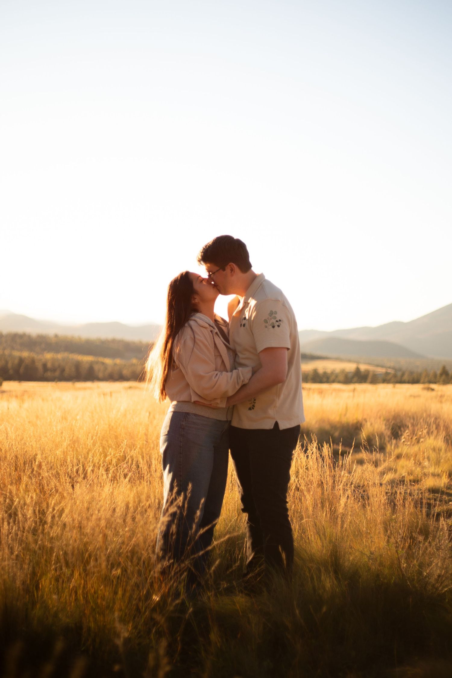 Couple kissing in a golden grass field at sunset with rolling mountains and a glowing sky in the background.
