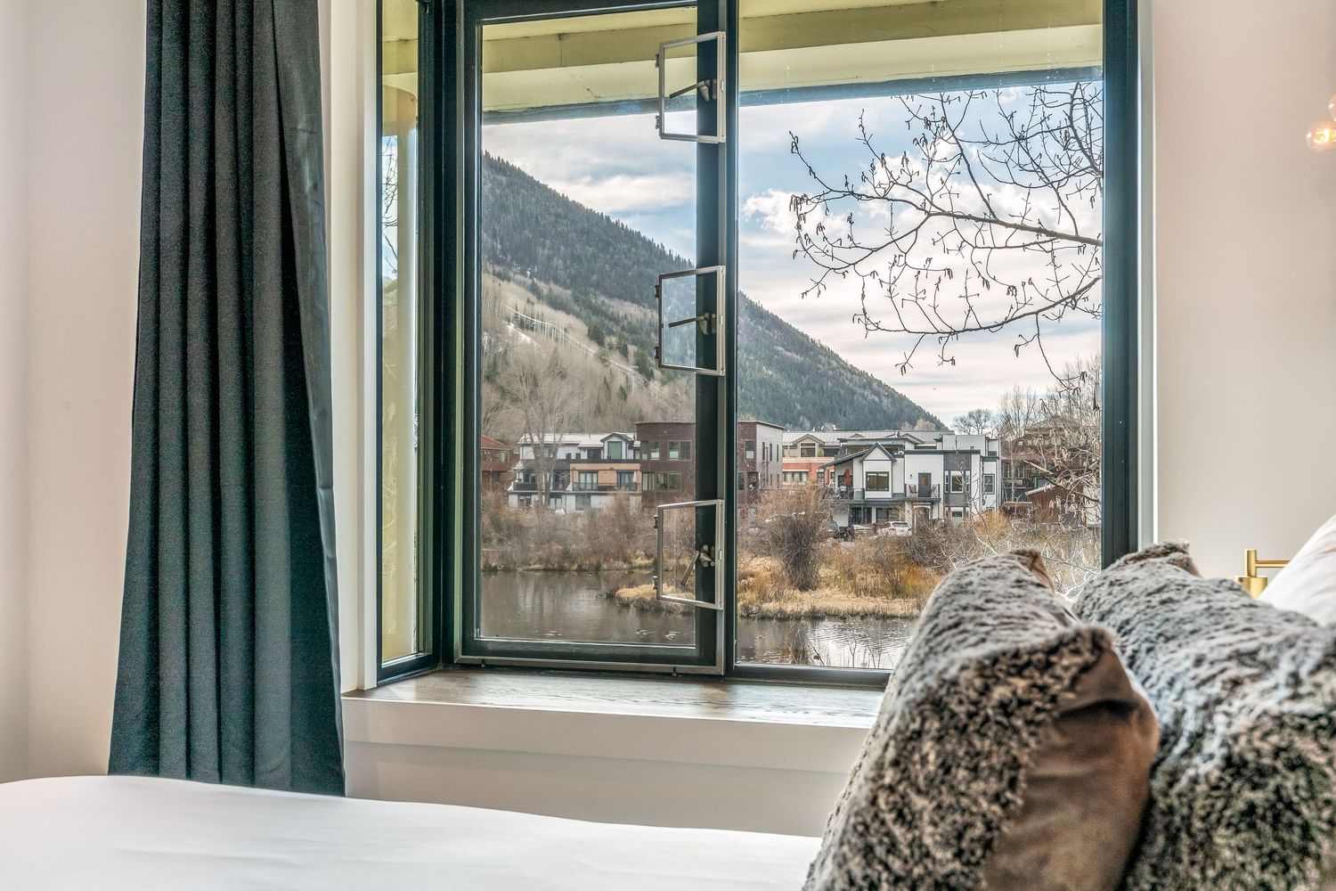 Bedroom window view of mountain town with river, surrounding peaks, and bare winter trees in foreground.