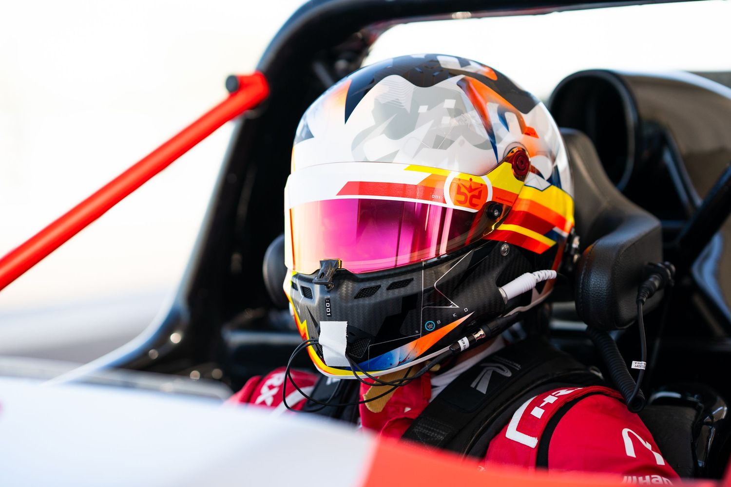 Racing driver in cockpit wearing colorful helmet with visor visible through windscreen.