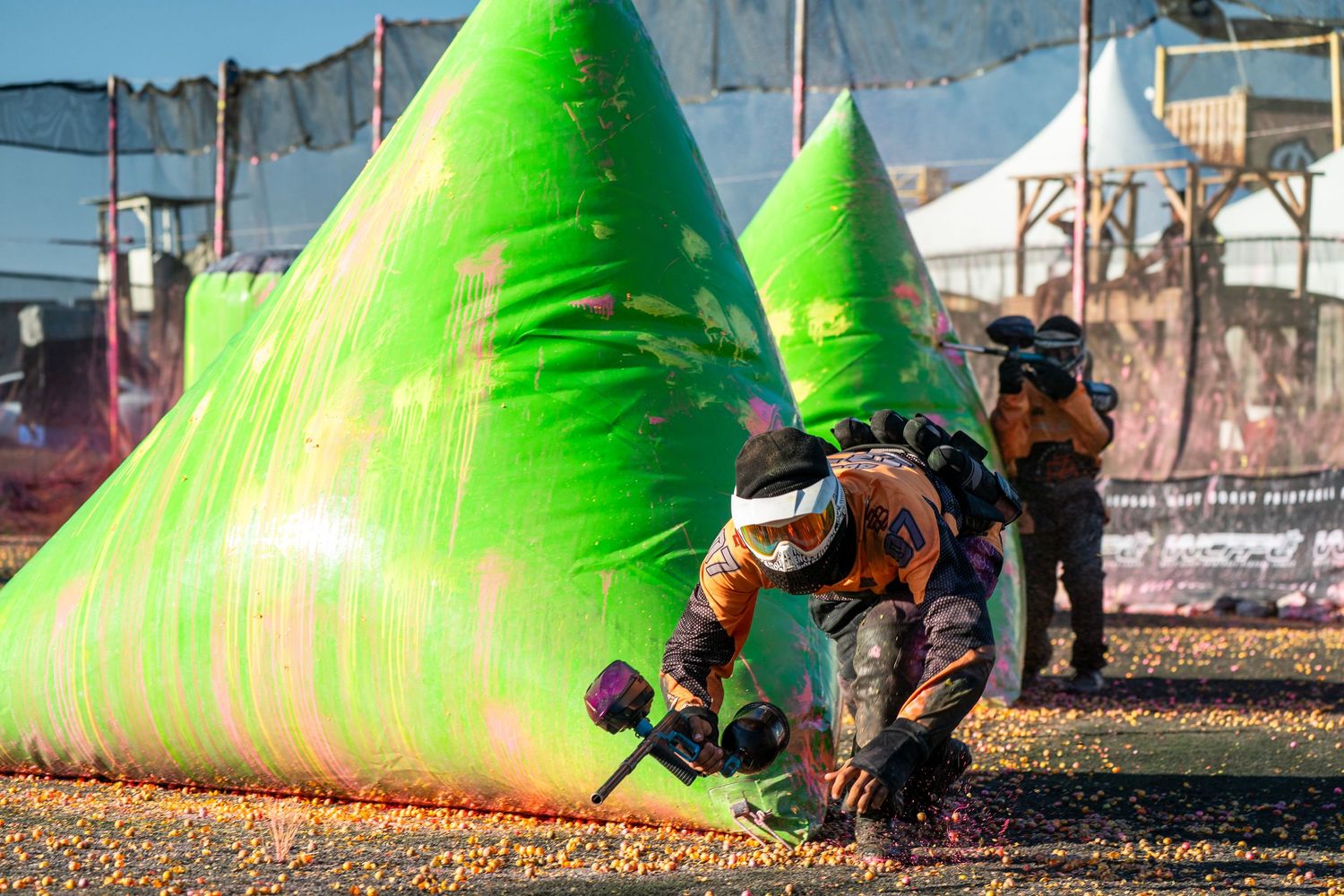 Paintball player crouching behind green inflatable bunker, aiming marker during outdoor tournament game.