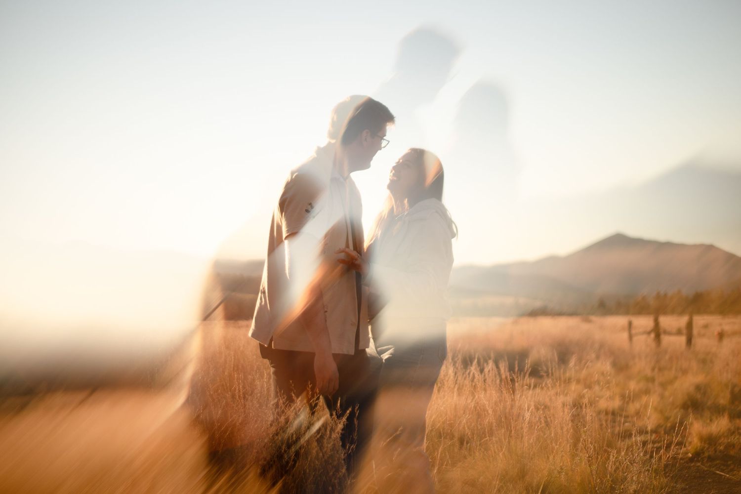 Double exposure photo of a couple embracing in a golden grassy field with soft silhouettes and distant mountains at sunset.