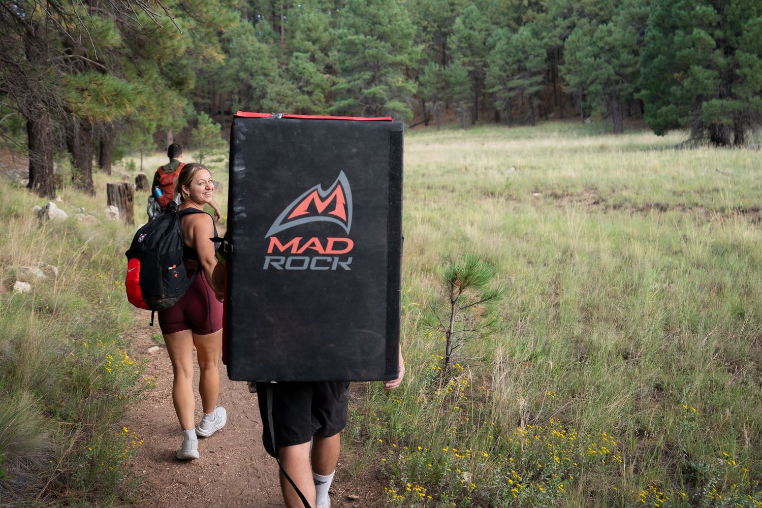 Two hikers carry Mad Rock crash pads and backpacks along a dirt trail through a sunlit grassy meadow toward a pine forest.