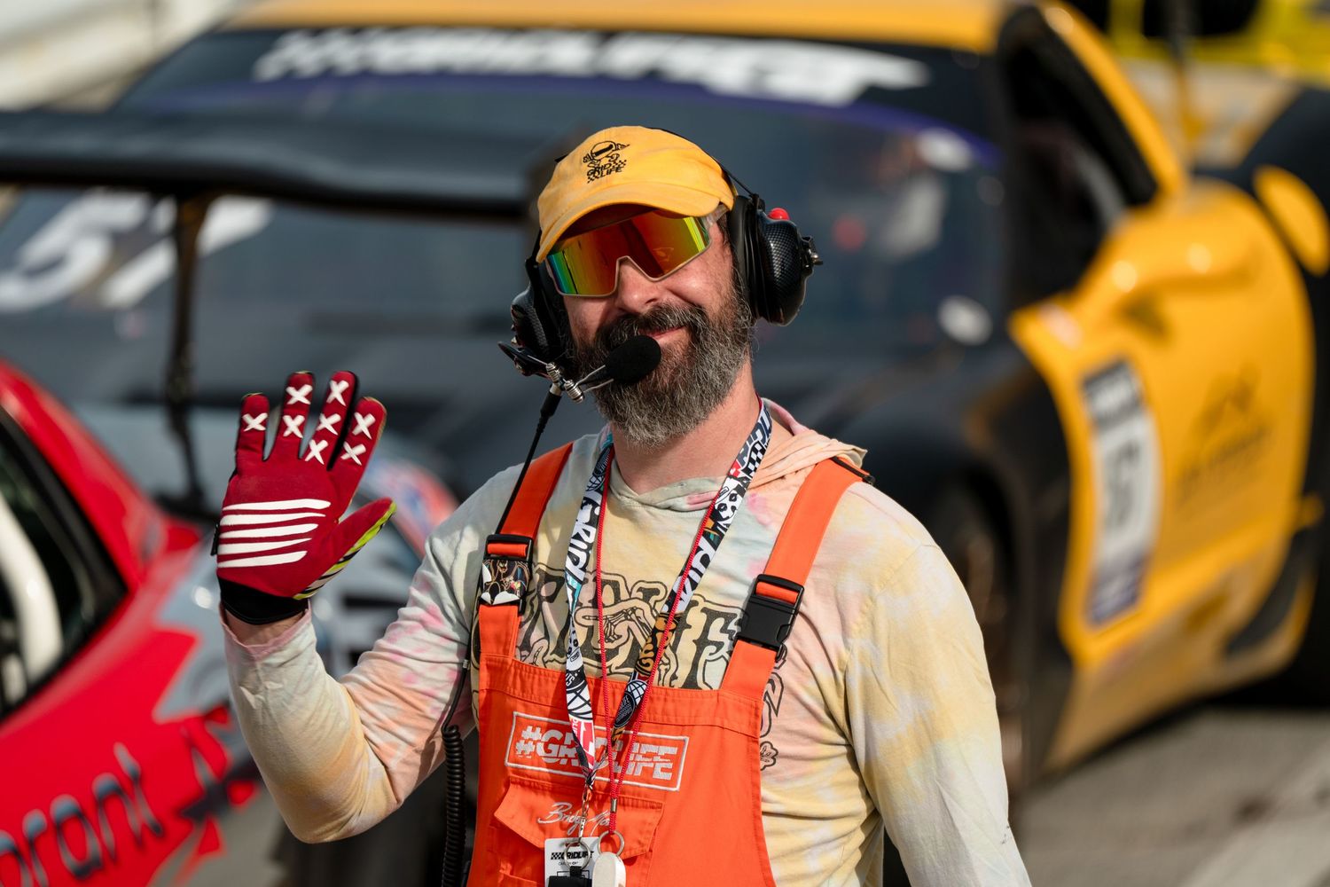 A bearded race official wearing headset and orange vest waves a flag at a motorsport event.
