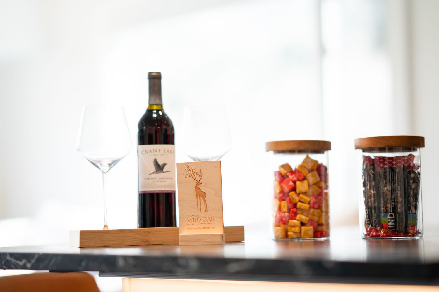 Red wine bottle with glass, wooden card holder, and two glass jars filled with colorful snacks on a dark counter.
