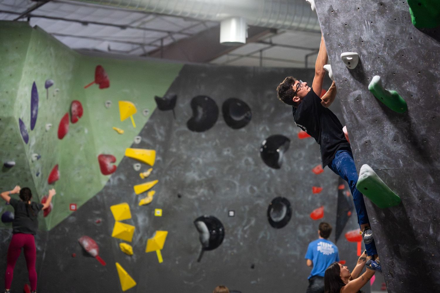 Climber in a black shirt and green climbing shoes hangs from an overhanging wall with colorful holds while others watch in a bouldering gym.