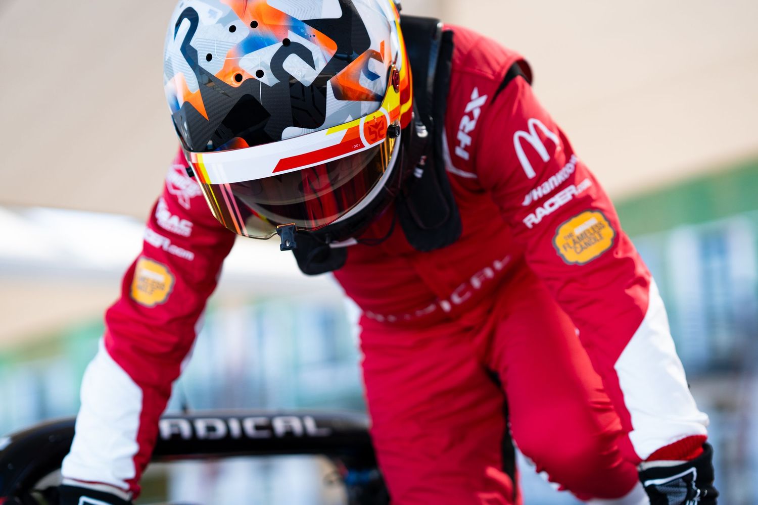 Racing driver in red suit with distinctive helmet design preparing in the cockpit.