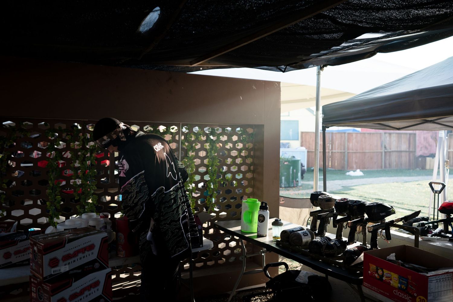 Person organizing tools and equipment under a shaded outdoor canopy area with green netting backdrop.