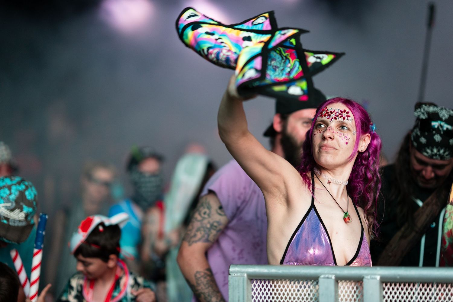 Festival goer with pink hair and glitter raises colorful hat at a lively music event.