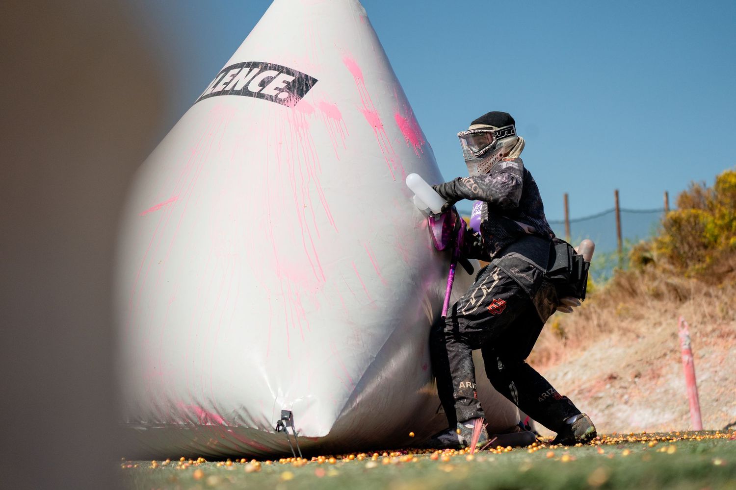 Paintball player in tactical gear crouches behind inflatable bunker during outdoor match.