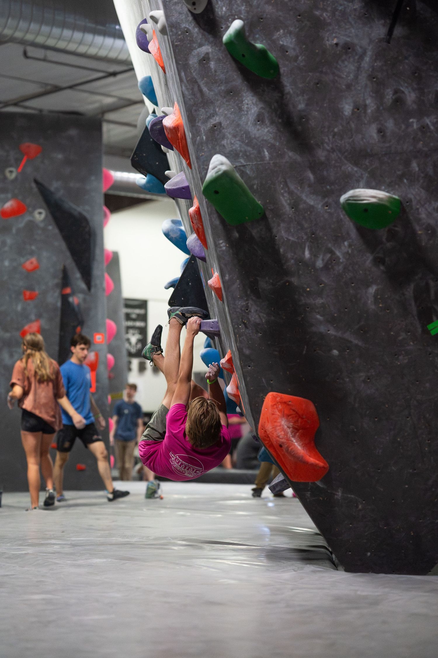Climber in a pink shirt scales an overhanging indoor bouldering wall while other climbers watch in a busy climbing gym.