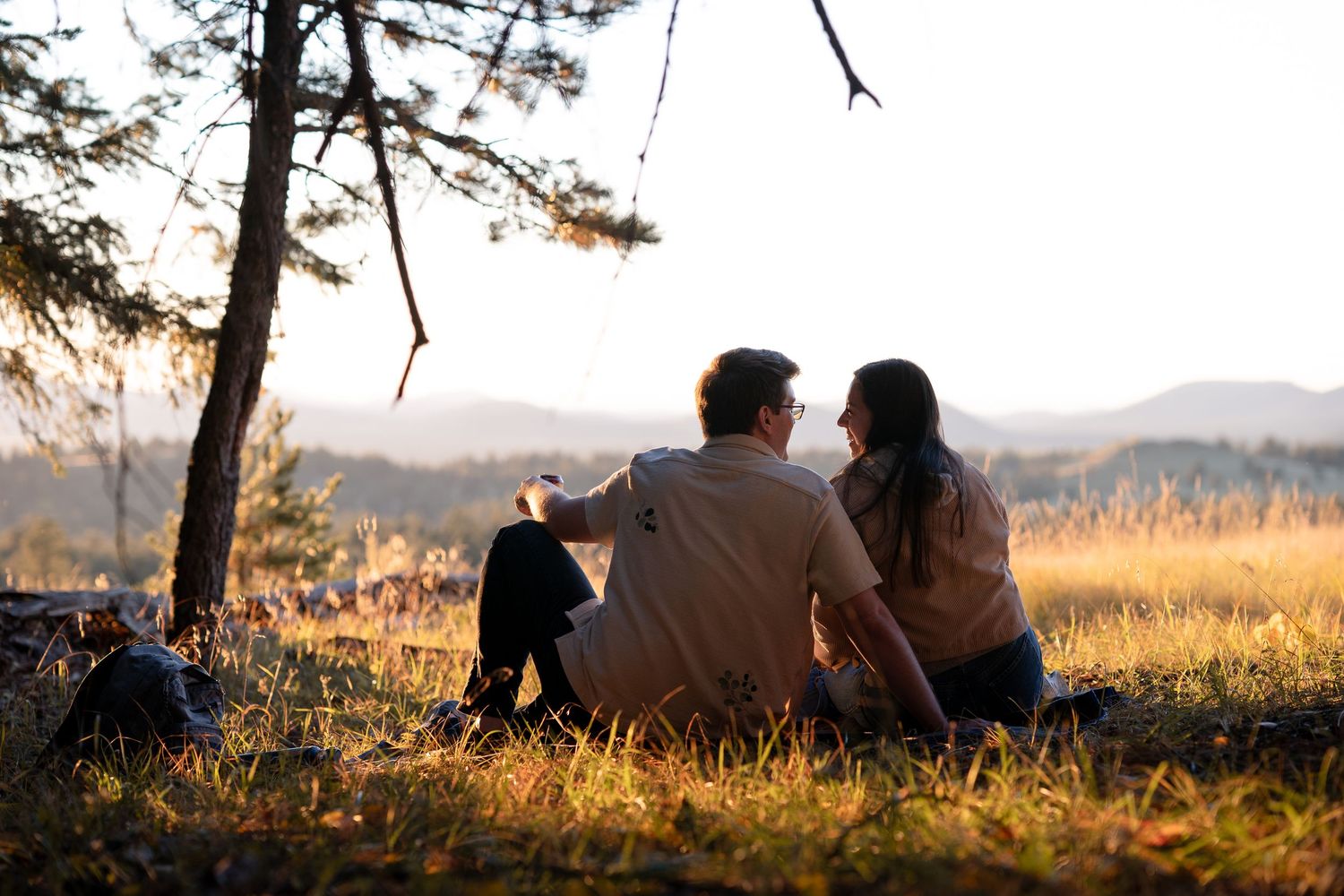 Couple sitting close together on a grassy hillside at golden hour, gazing out at distant mountains beside a pine tree.