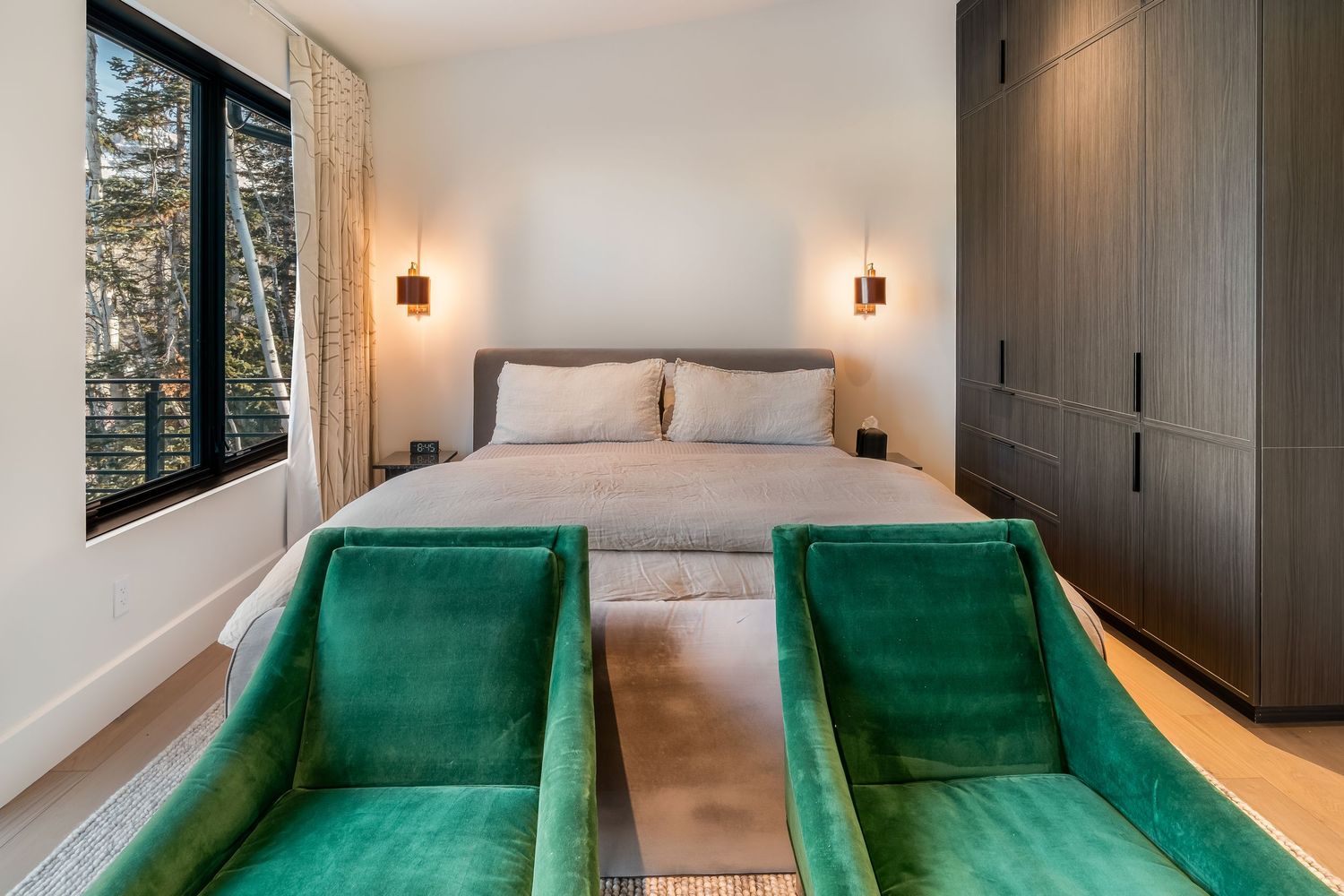 Modern bedroom with emerald green chairs and built-in storage features a neutral color scheme and wall sconces.
