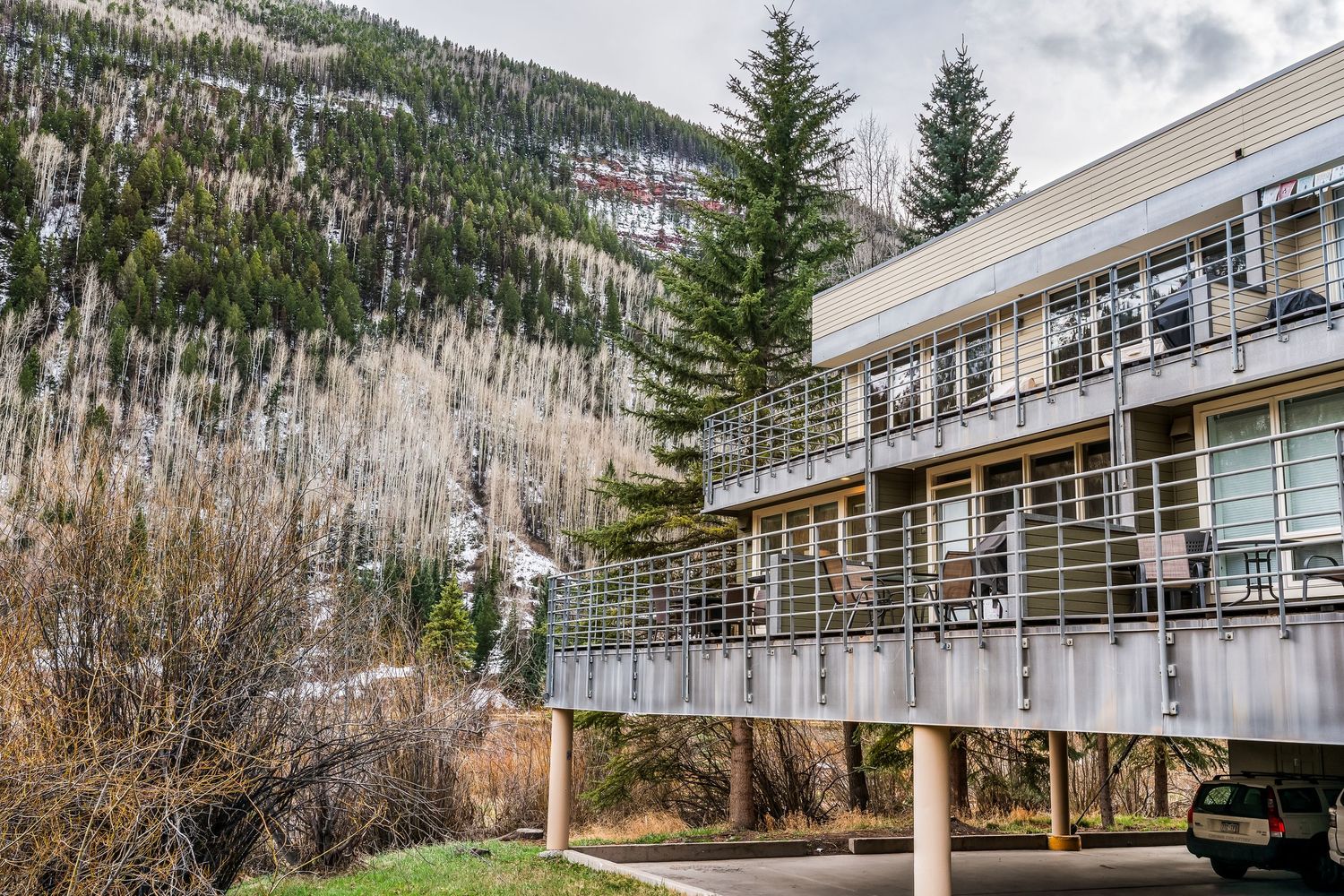 Multi-story mountain lodge with balconies nestled against a forested hillside in a scenic alpine setting.
