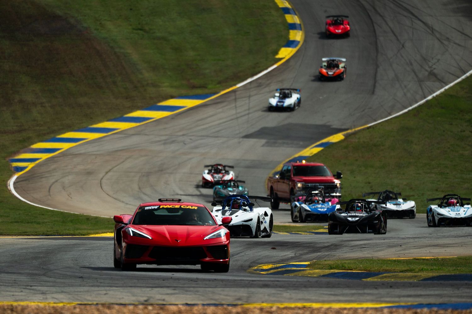 Red Corvette pace car leads a pack of racing cars through a curve at a professional road course circuit.