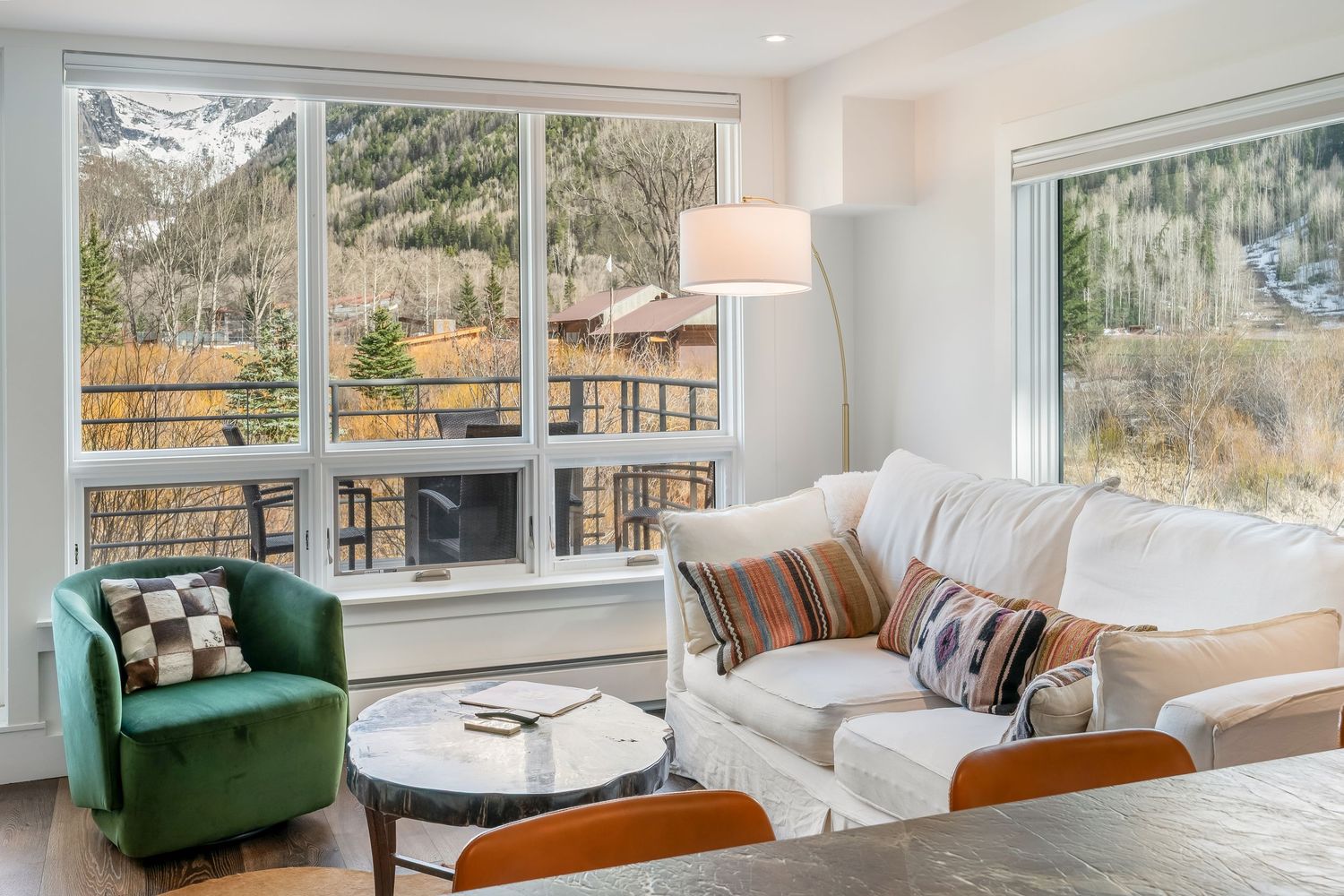Modern mountain condo living room with white sofa, green velvet chair, and panoramic windows overlooking autumn forest views.