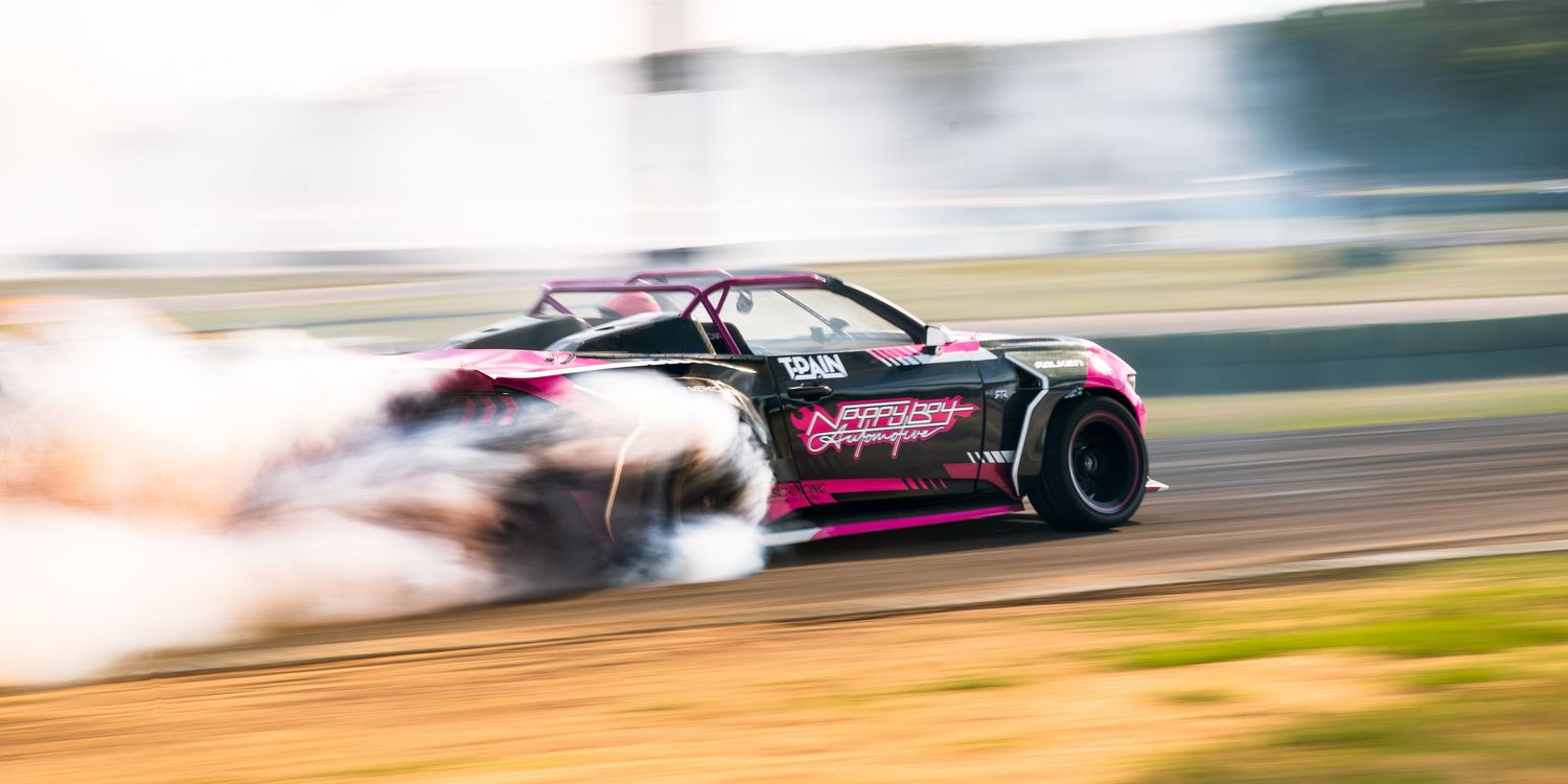 Pink and black drift car spinning with smoke on a dirt track during a high-speed motorsport competition.