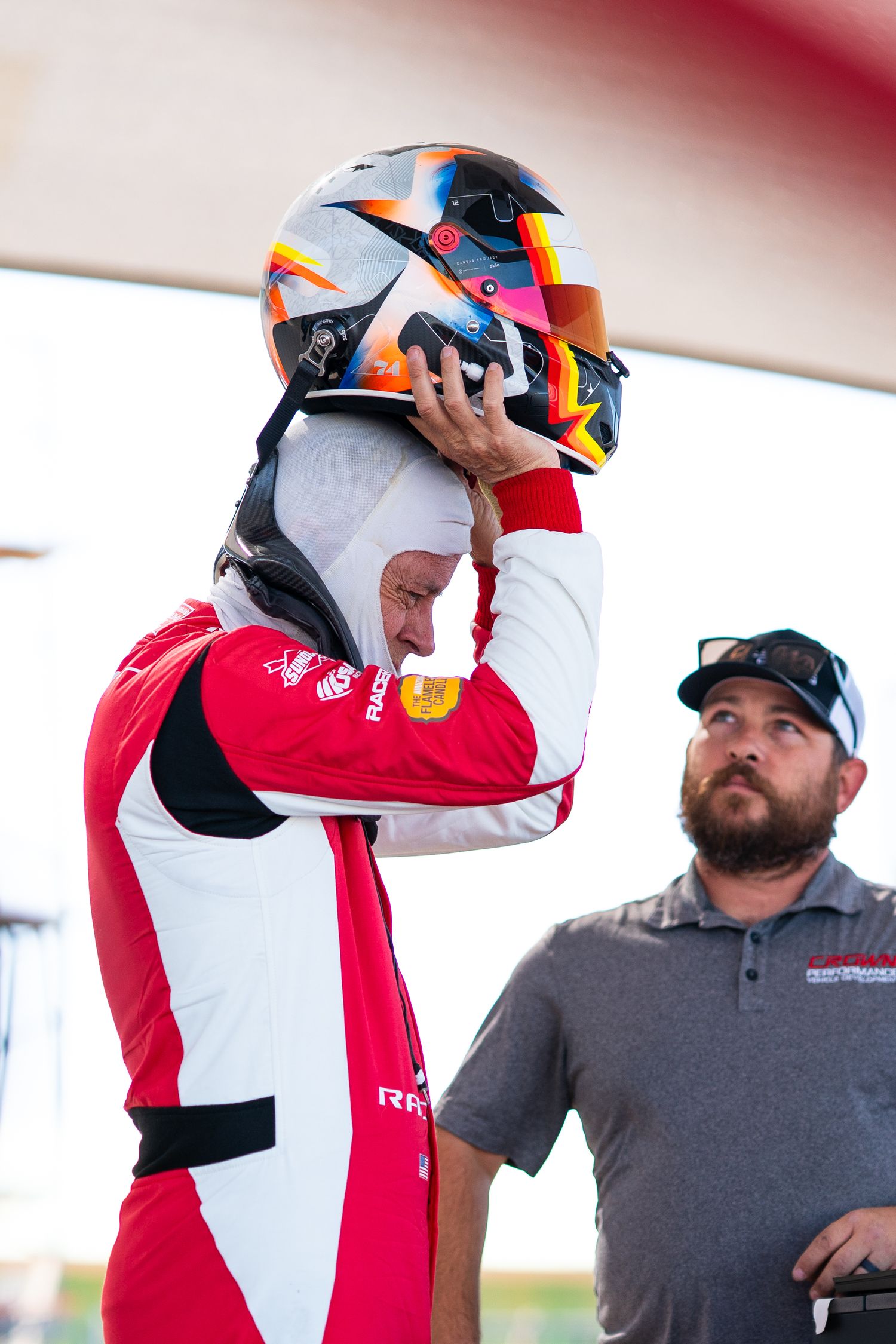 Racing driver in red and white suit adjusting helmet while crew member looks on.