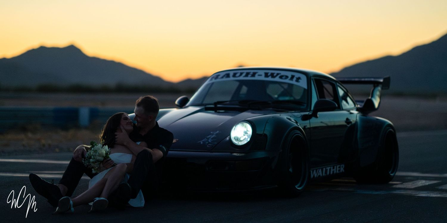 Black Porsche 911 with wide body kit parked at sunset against mountain landscape.