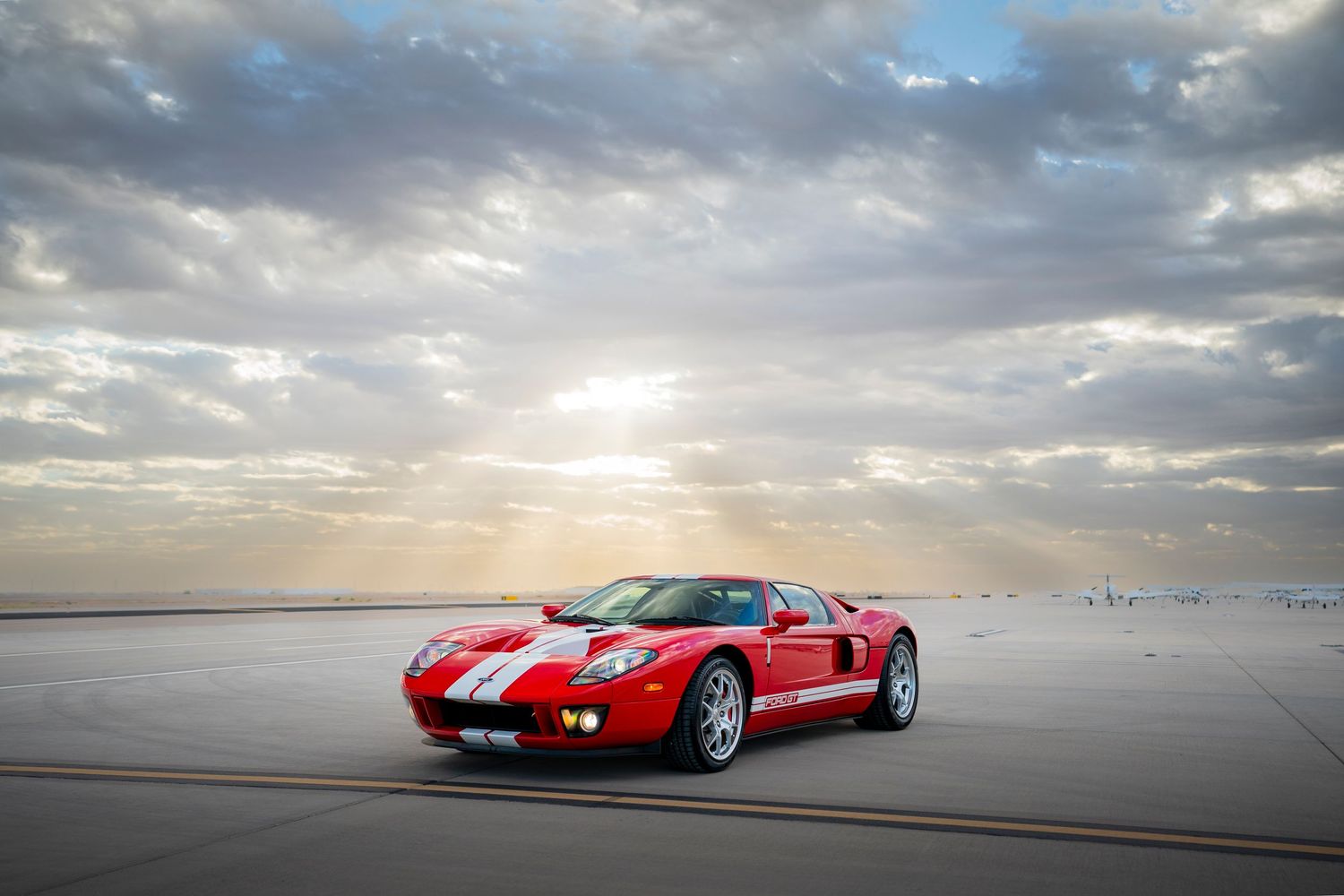 Red Ford GT supercar with white stripes parked on airfield under dramatic cloudy sunset.