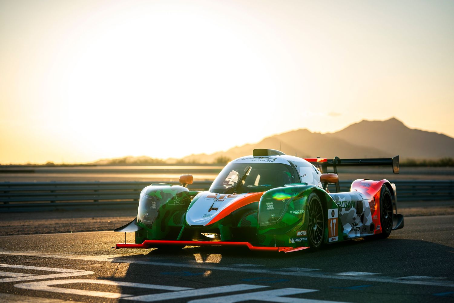 Green and red racing prototype car on track during golden sunset with mountains in background.