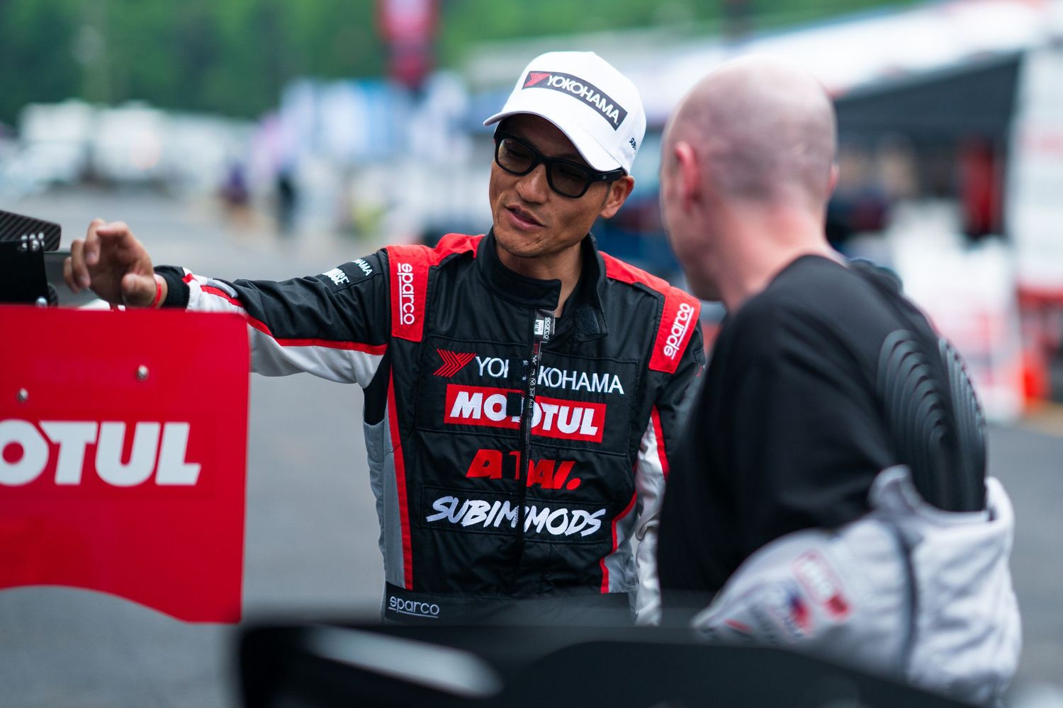 Racing driver in Motul Subaru gear conversing with crew member at motorsport event.