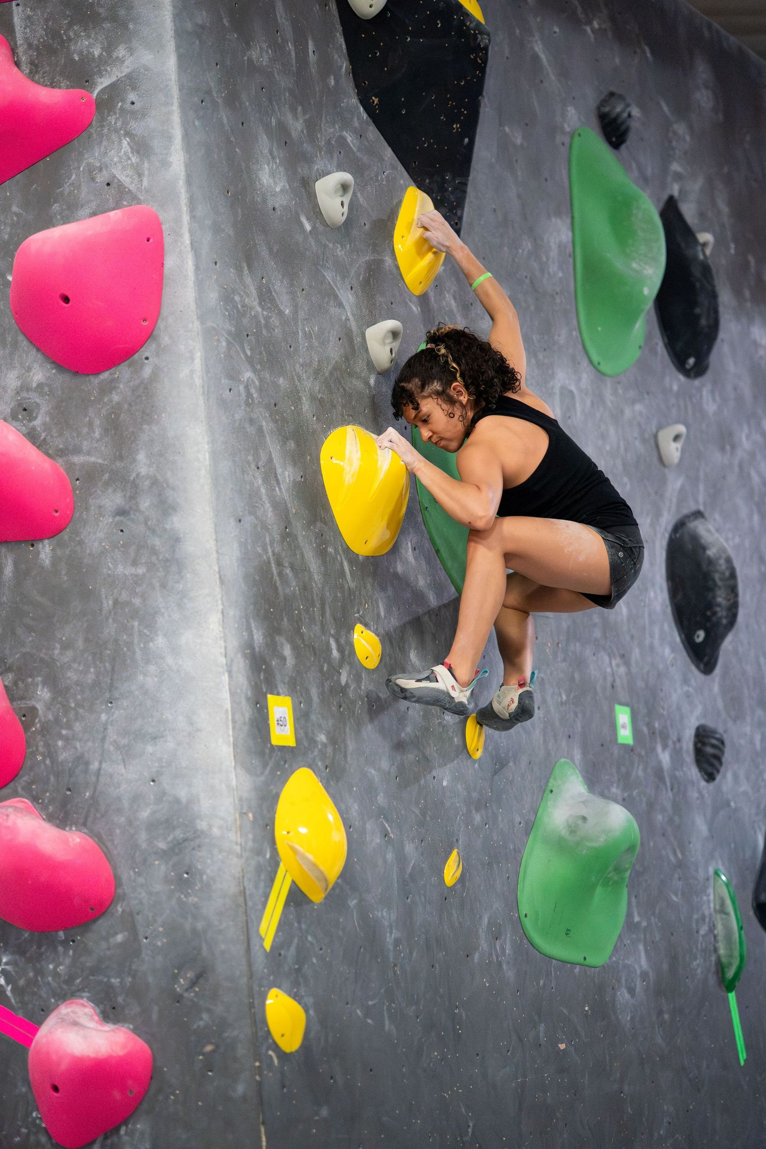 Climber in a black tank top and shorts reaches for a yellow hold on a gray indoor bouldering wall with colorful pink and green holds.