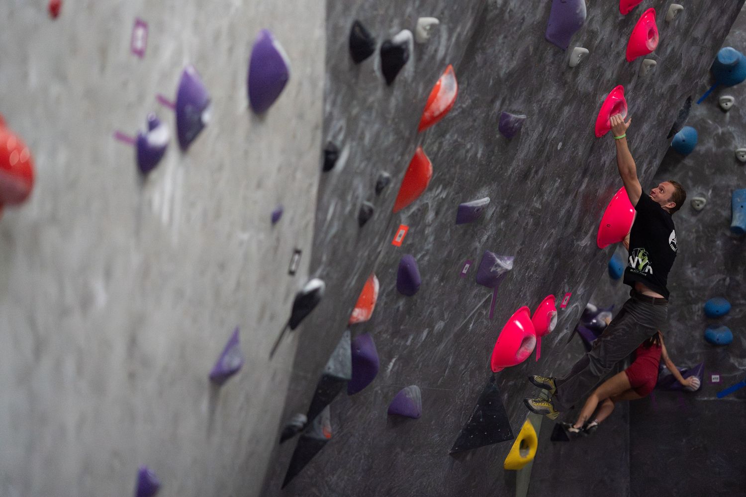 Climber in a black shirt and red shorts reaching for a hold on an indoor bouldering wall covered in colorful purple, red and orange holds.