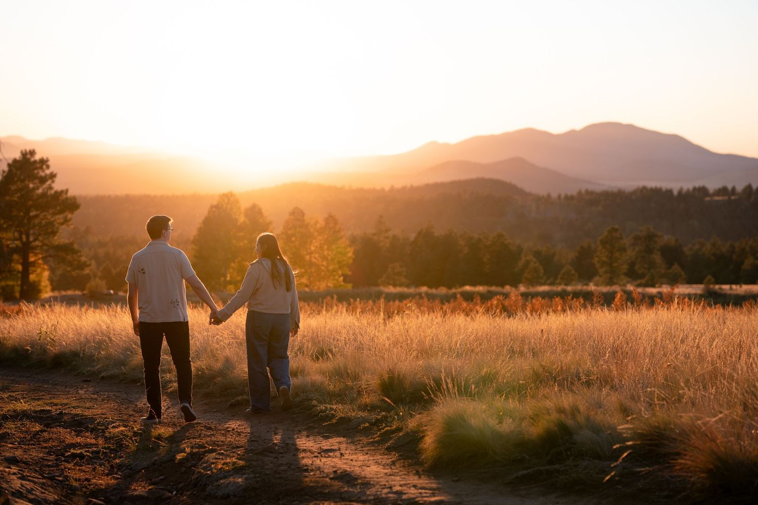 Couple holding hands walking along a dirt path through golden grass at sunset with mountains and pine trees in the background.