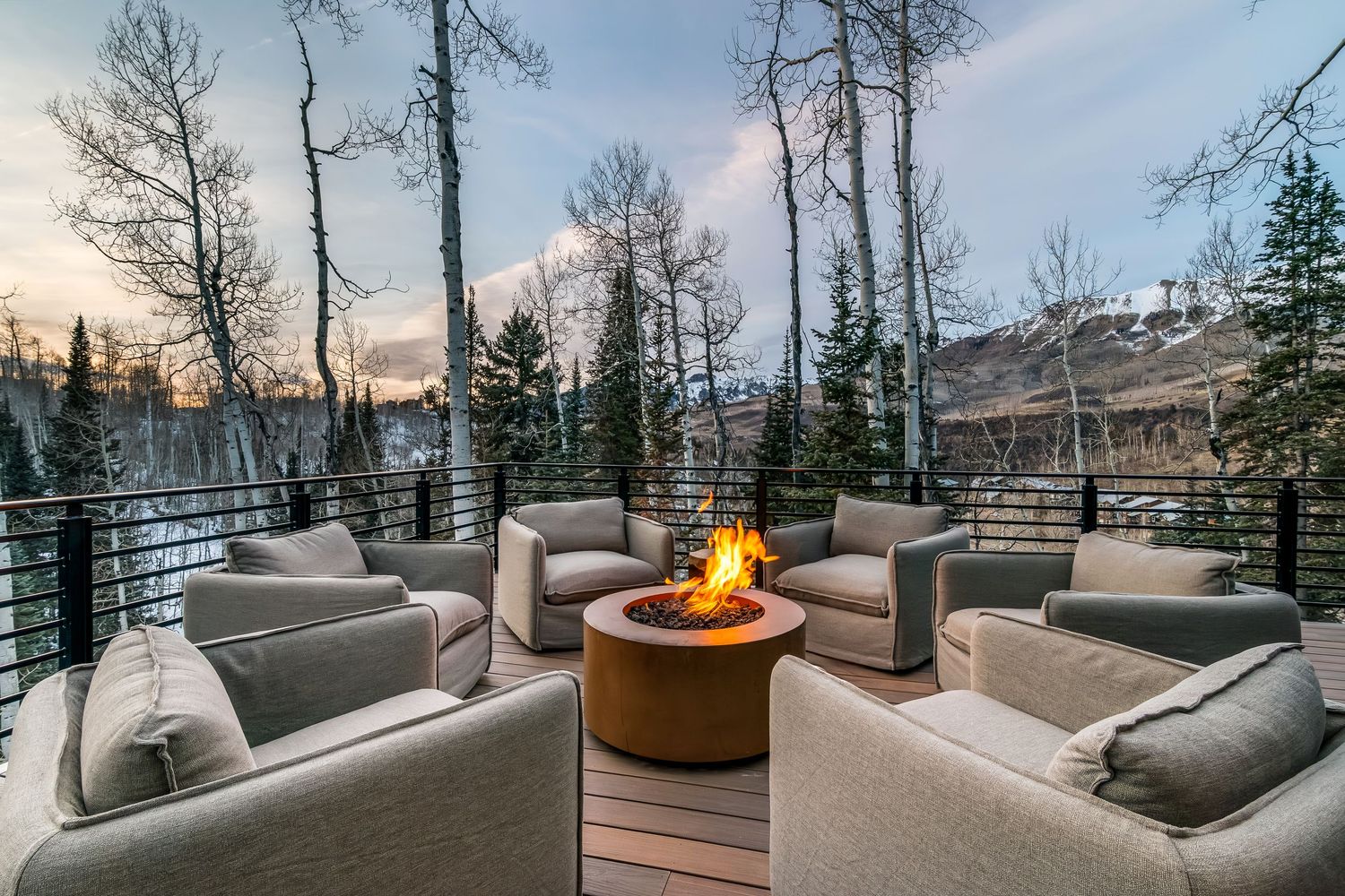Luxurious mountain lodge deck with outdoor firepit and panoramic views of snowy peaks at sunset.