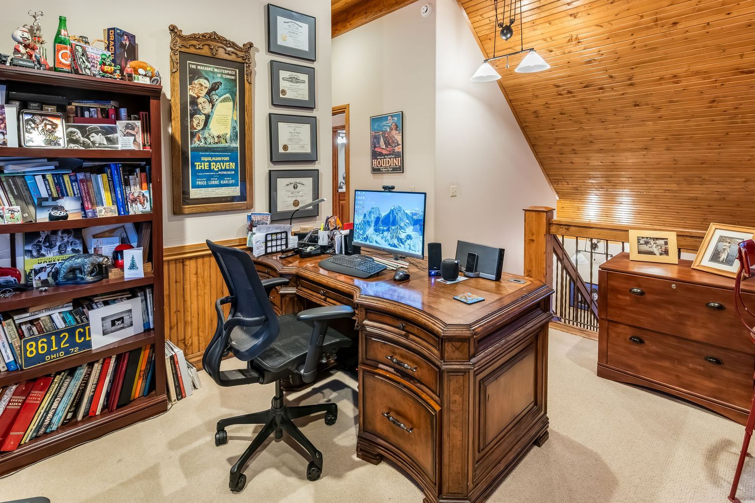 Home office with wooden desk, bookshelf, and pine ceiling featuring rustic decor and mountain artwork.