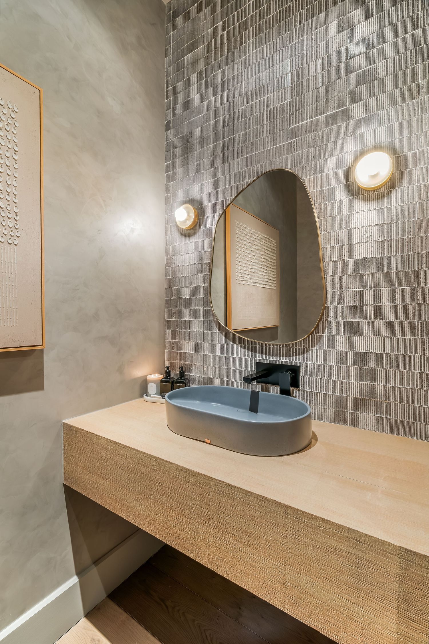 Modern bathroom vanity with round mirror, wooden countertop, and gray textured tile walls illuminated by wall sconces.