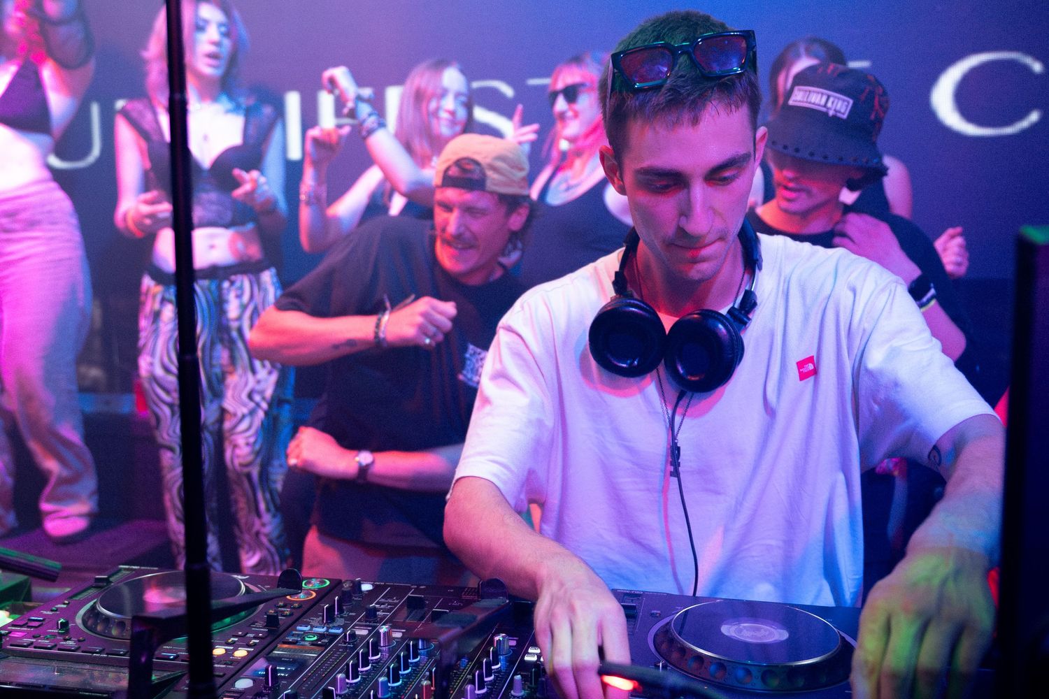 DJ performs at a nightclub with headphones around neck while working mixer deck in purple-lit atmosphere.