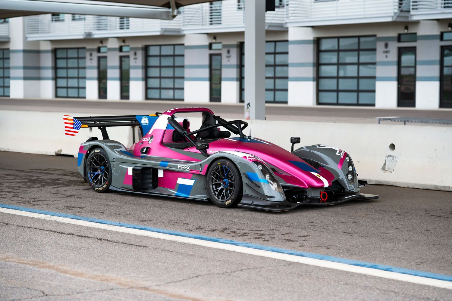 Pink and black racing prototype car with large rear wing drives on racetrack past pit wall.
