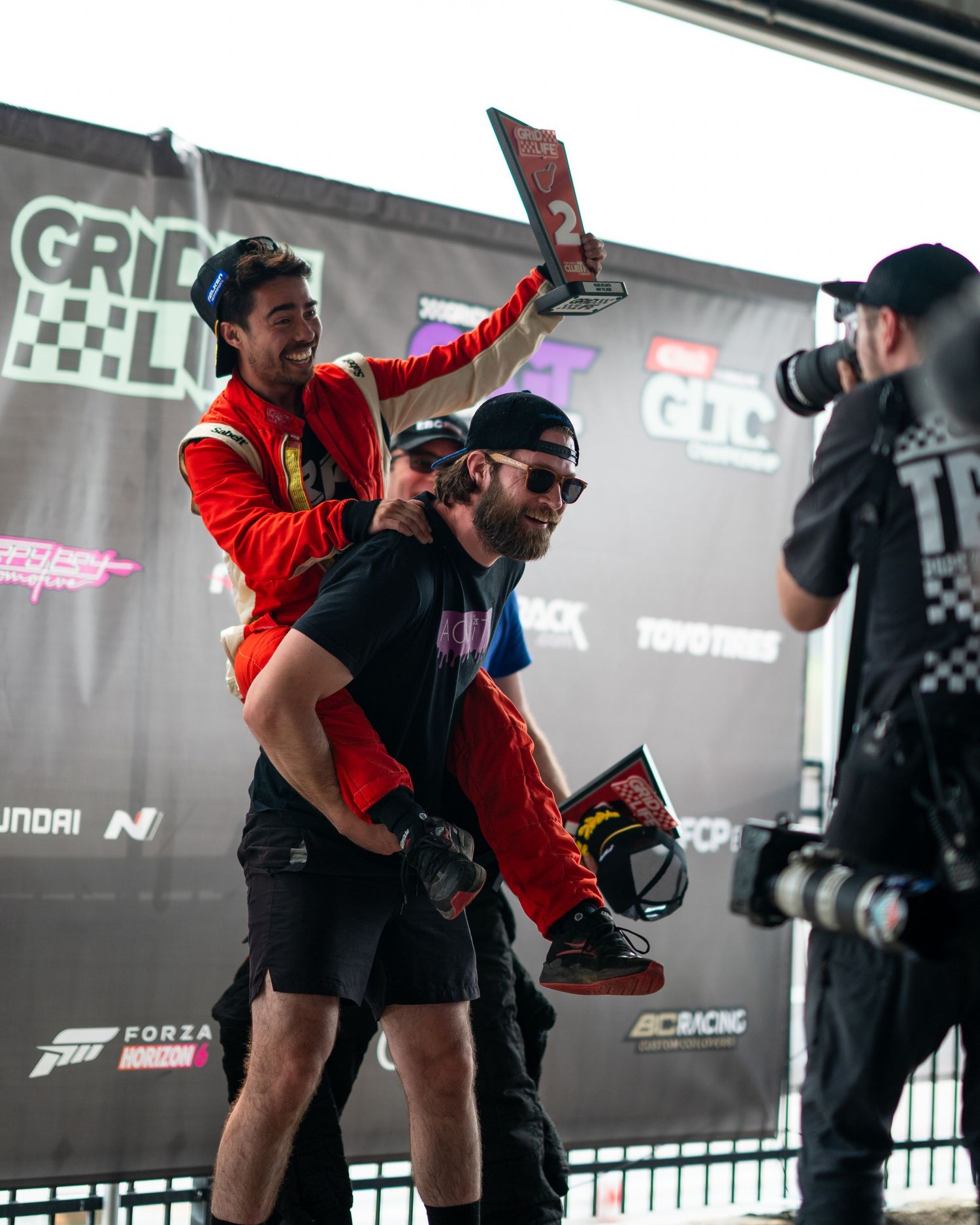 Excited racing driver celebrates victory on teammate shoulders at Gridlife motorsport event podium.