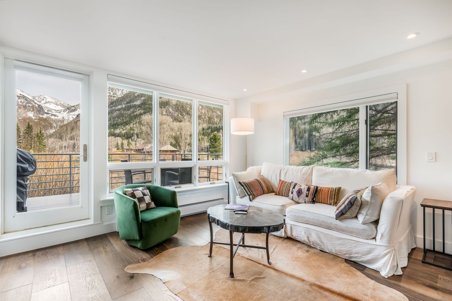 Bright living room with white sofa, green velvet chair, cowhide rug, and large windows overlooking snowy mountain views.