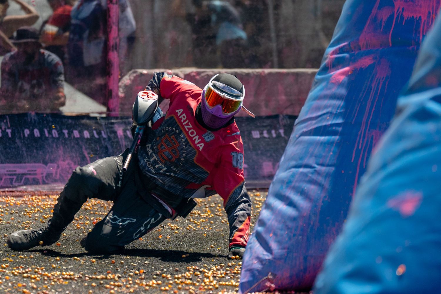 Paintball player in red gear crouching behind inflatable bunker during competitive paintball tournament match.