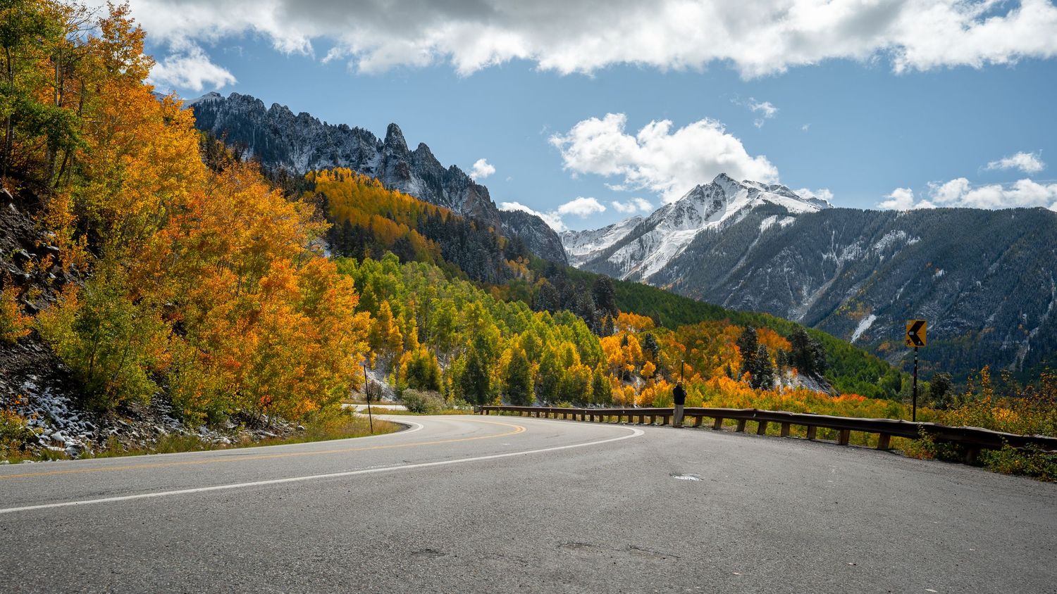 Winding mountain road lined with golden aspen trees and snow-capped peaks in the distance.