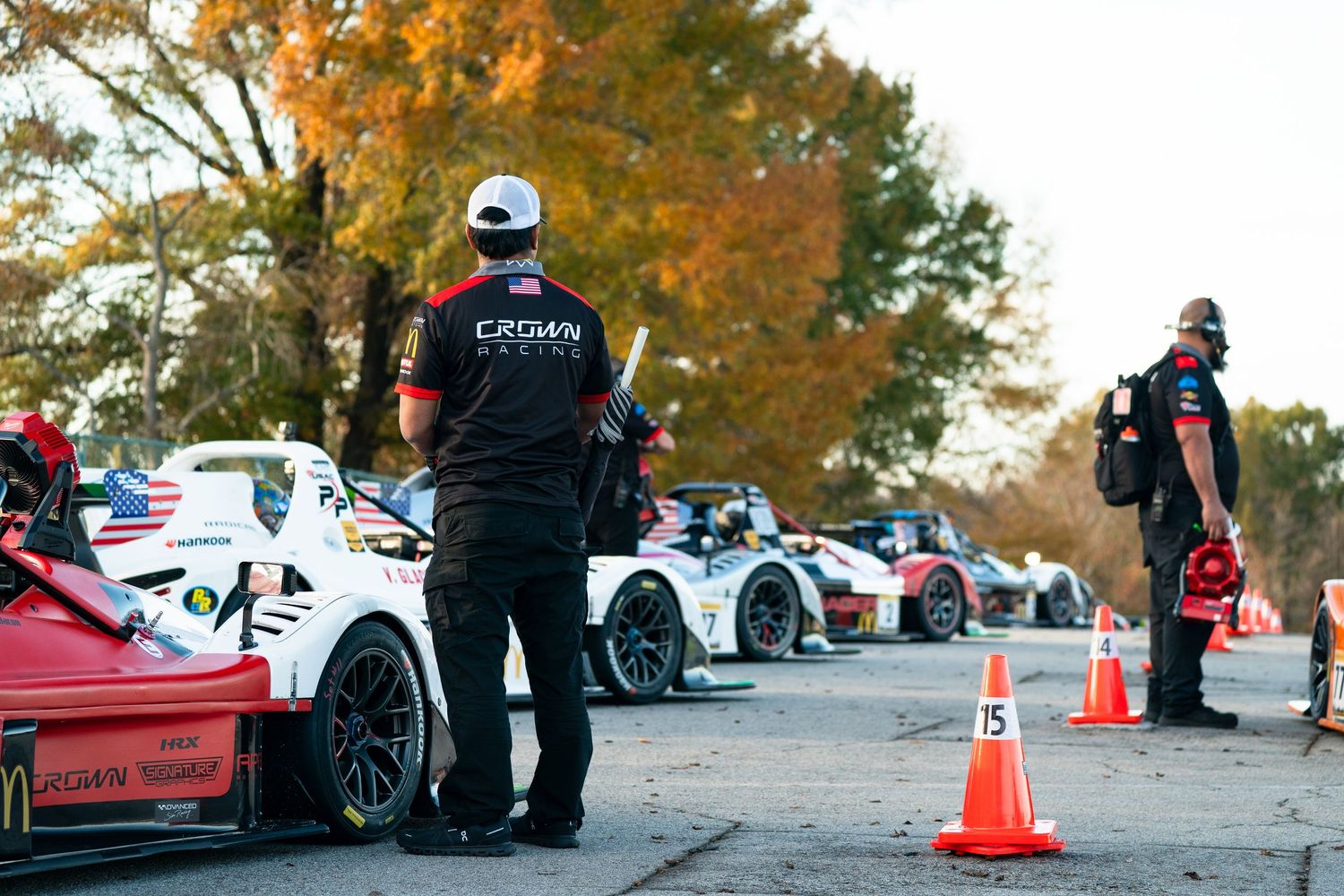 Row of cars parked at an outdoor event with orange autumn trees and traffic cones in the background.